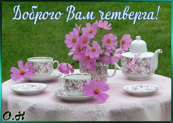 Уютное чаепитие с розовыми цветами и фарфором. Приветственная открытка «Доброго Вам чемберга!» для создания теплого настроения и весеннего уюта. Открытка Доброго Четверга! Уютная и светлая композиция с чашкой чая и нежными розовыми цветами космеи на фоне зеленой травы. На столе накрыта белая кружевная скатерть, стоит изящный фарфоровый чайник и чашки с цветочным принтом. На изображении красуется приветственная надпись на украинском языке «Доброго Вам чемберга!», создающая атмосферу тепла, домашнего уюта и весеннего настроения