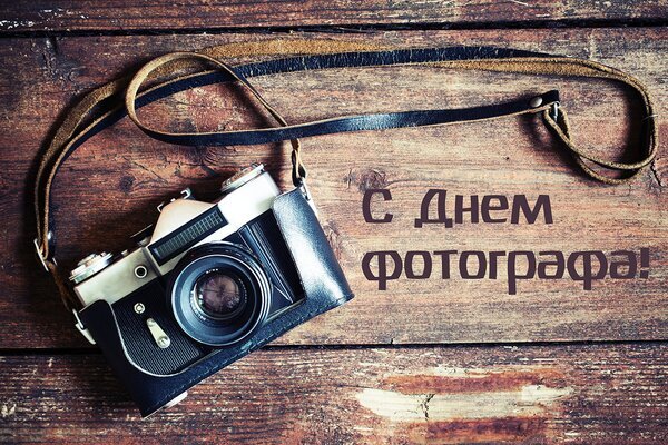 Поздравление с Днем фотографа: винтажный фотоаппарат на деревянном столе и праздничная надпись. Идеально для открытки и соцсетей. Открытка С Днем фотографа! Поздравительная открытка к Дню фотографа. На деревянном фоне лежит винтажный пленочный фотоаппарат с кожаным ремешком. Справа расположена стильная надпись на русском языке «С Днем фотографа!»