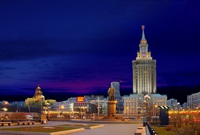 Ночная Москва: величественный сталинский небоскреб и городские огни в глубоких синих сумерках. Панорамный вид на архитектуру мегаполиса. Открытка ночной город москва Вечерний панорамный вид на величественную архитектуру Москвы в сумерках. На переднем плане запечатлен монументальный сталинский небоскреб, ярко освещенный огнями, на фоне глубокого темно-синего ночного неба. В композиции также видны городские огни, памятник на постаменте и элементы городской инфраструктуры, создающие атмосферу торжественного и спокойного мегаполиса