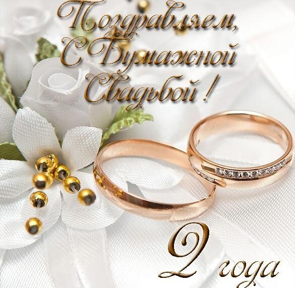 Поздравление с 2-й годовщиной свадьбы (Бумажная свадьба). Красивое фото с золотыми кольцами и белыми розами для праздничной открытки. Открытка С бумажной свадьбой 2 года Эстетичное поздравительное изображение к годовщине свадьбы. На светлом фоне с белыми розами крупным планом изображены два золотых обручальных кольца. В верхней части кадра стильным золотистым шрифтом нанесена надпись на русском языке: «Поздравляем с Бумажной Свадьбой!», а внизу — «2 года»